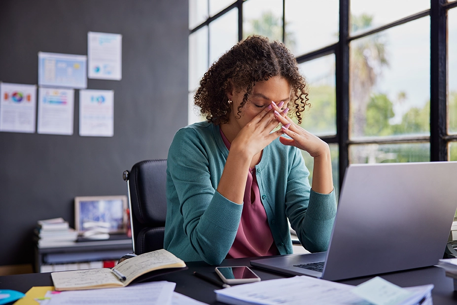 Person sitting at a desk with eyes closed and hands near face, looking stressed while working on a laptop in an office with papers, notebook and phone nearby